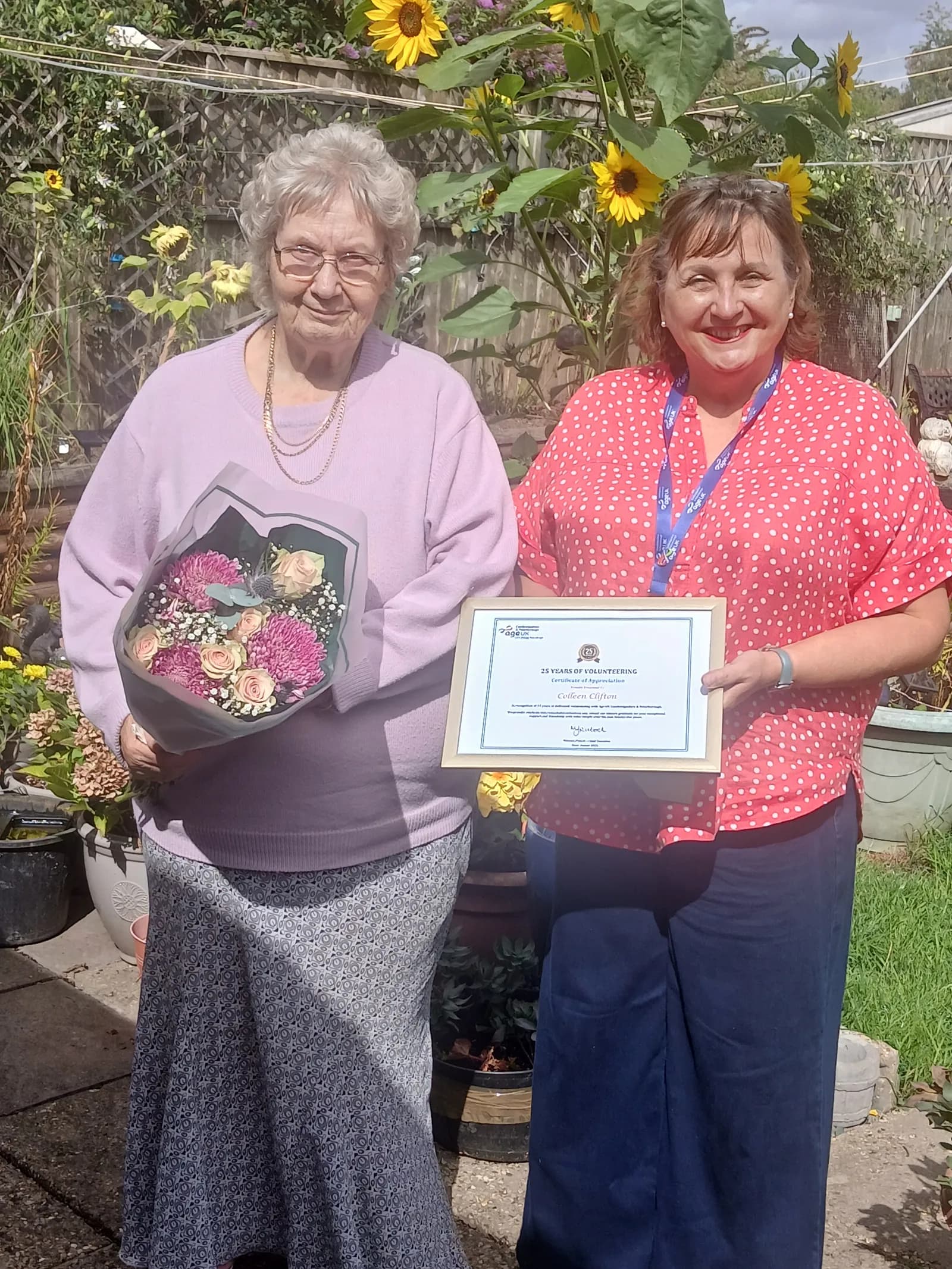 Two women stand in a garden with sunflowers, one holding a bouquet of flowers and the other holding a framed certificate.